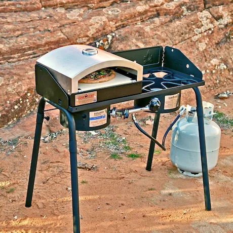 Ein tragbarer Outdoor-Pizzaofen, der in einer felsigen Umgebung aufgestellt ist. Der Ofen ist mit einem Propantank verbunden, und eine Pizza wird darin gebacken.